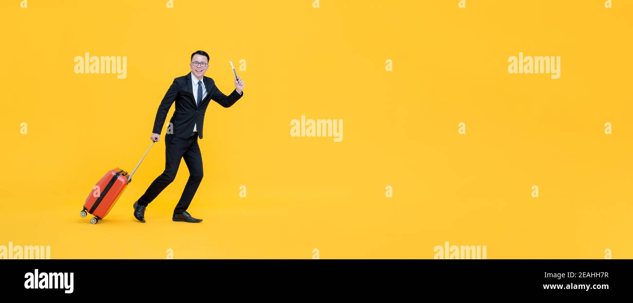 Full body of Asian man in formal business suit with baggae passport and boarding pass ready to go for traveling isolated on yellow banner background Stock Photo
