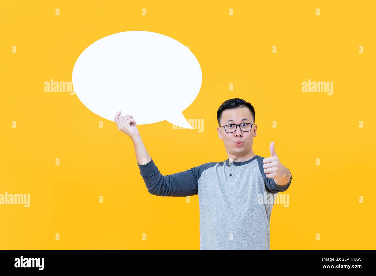 Excited shocked young Asian man giving thumbs up while holding blank speech bubble Stock Photo