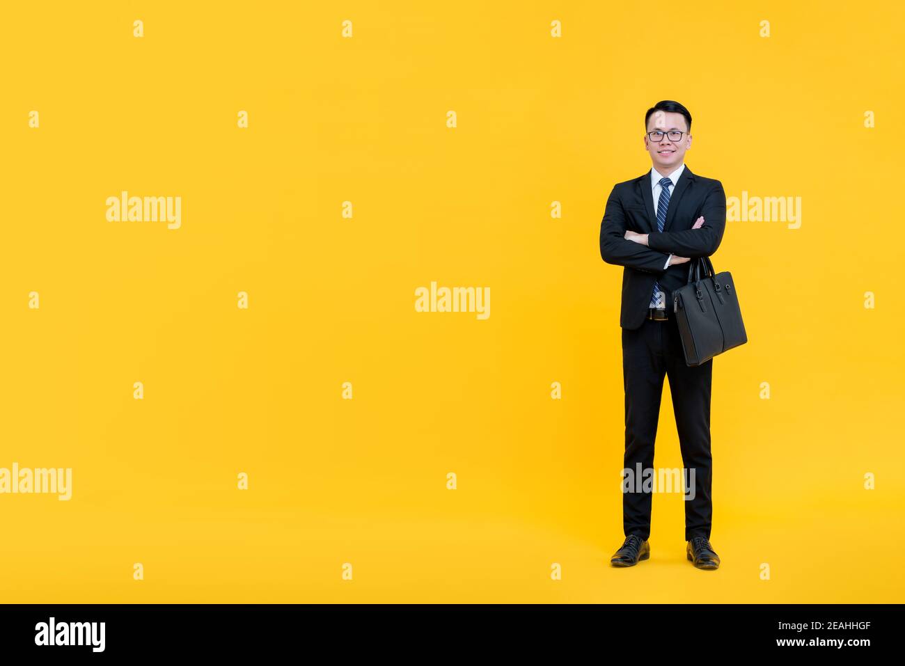Asian businessman in formal business suit standing with arms crossed holding briefcase isolated on yellow background with copy space Stock Photo