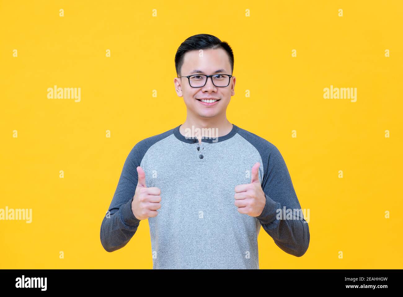 Optimistic Asian man in casual t-shirt smiling giving thumbs up isolated on yellow background Stock Photo