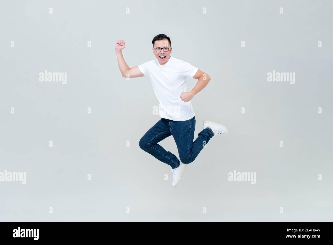 Energetic Asian man in casual white t-shirt and jeans jumping on light gray background Stock Photo
