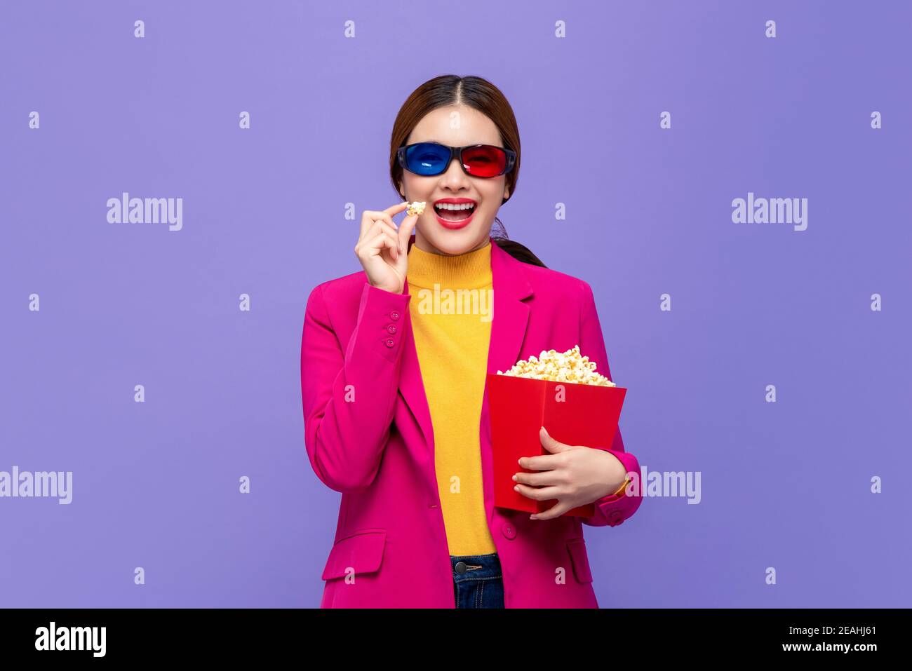 Young Asian woman wearing 3D glasses enjoying eating popcorn and watching movie isolated on purple background Stock Photo
