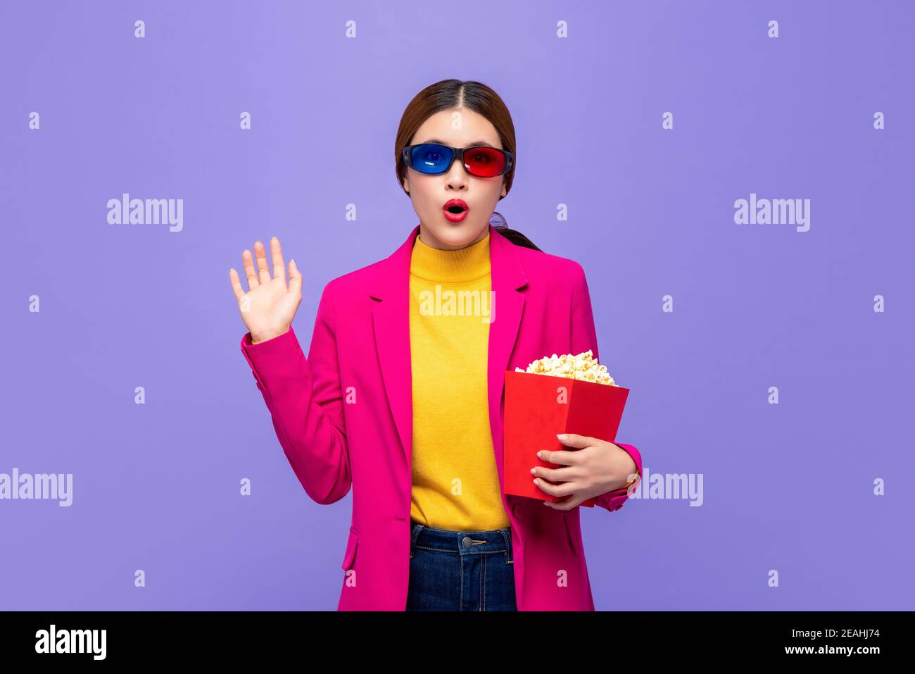 Excited Asian girl in colorful clothes with 3D glasses holding popcorn and wacthing movie  isolated on purple background Stock Photo