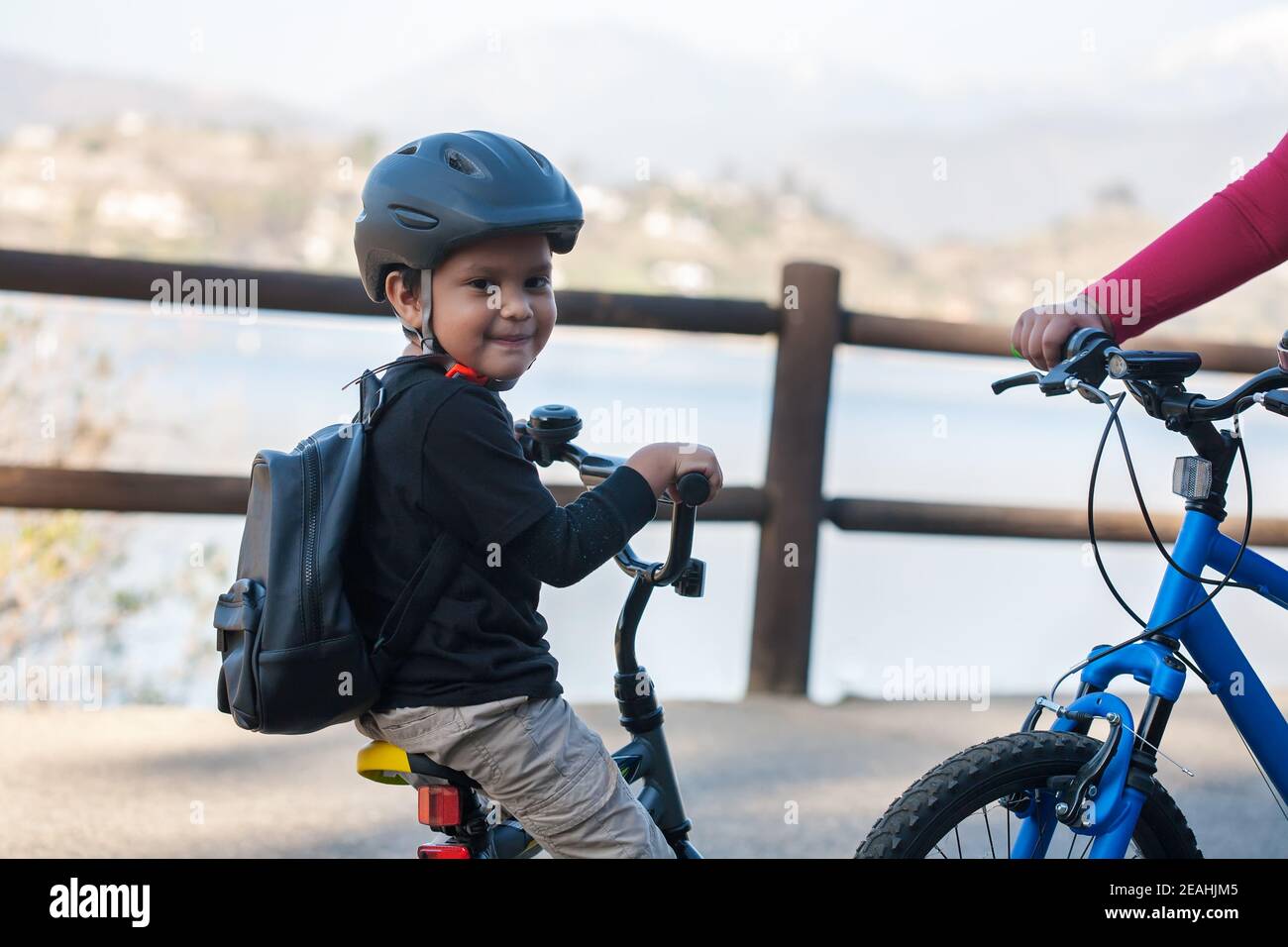 A Latino boy wearing a backpack and helmet on a small kids bike, learning to ride a bicycle by a lake and standing next to a larger mountain bike. Stock Photo