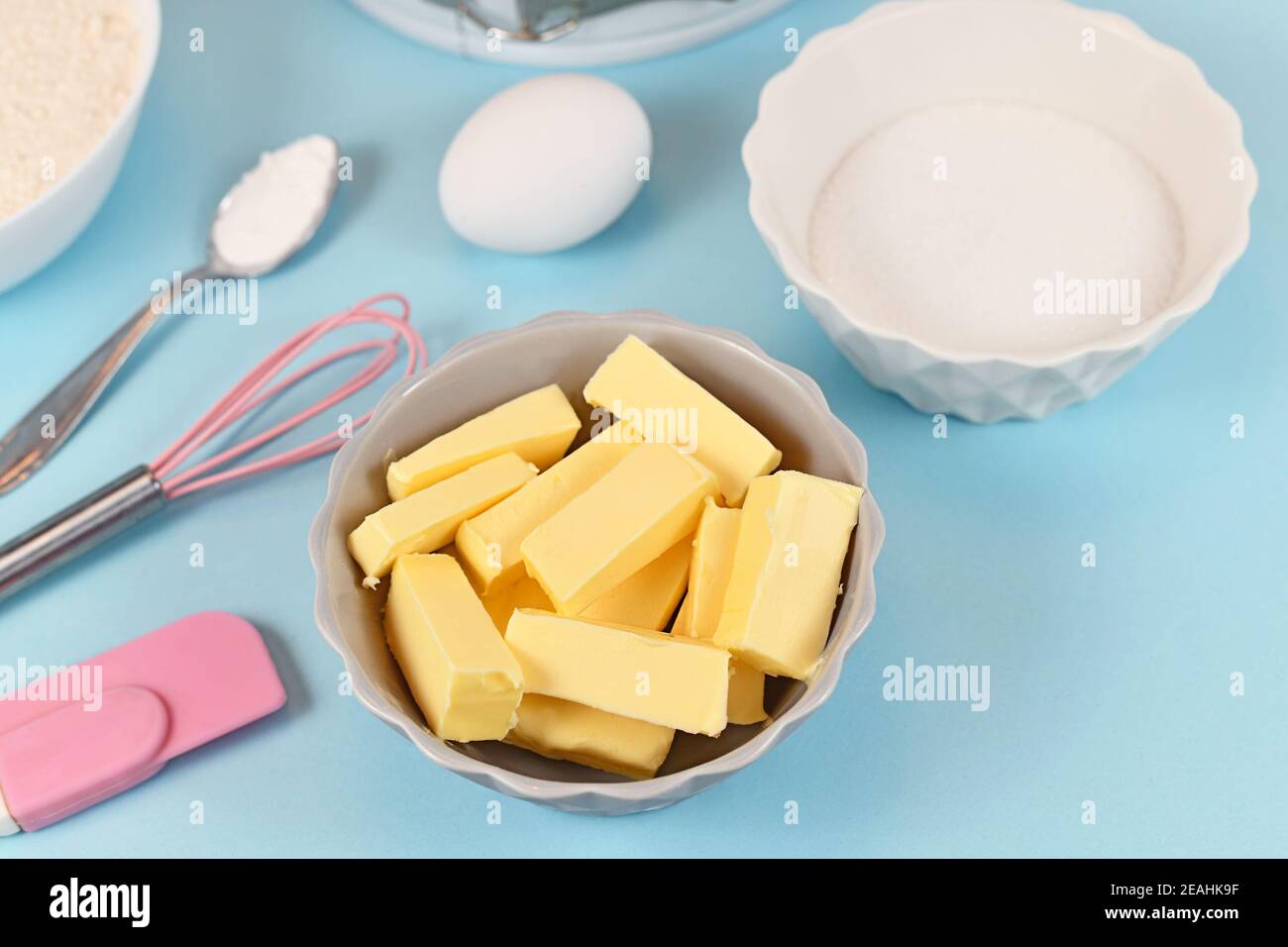 Butter cut in pieces for preparing dough for shortcrust cake base with sugar, egg, baking bowder and baking utensils on blue background Stock Photo