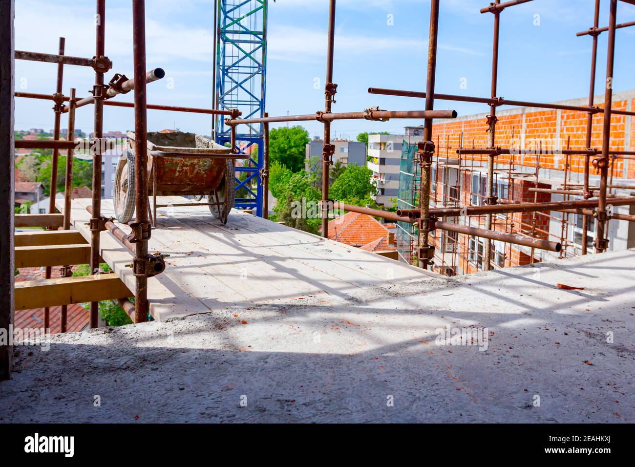 Used industrial obsolete wheelbarrow placed on wooden platform above building site as a part of scaffold placed against edifice under construction for Stock Photo