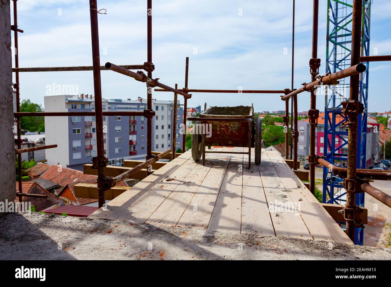 Used industrial obsolete wheelbarrow placed on wooden platform above building site as a part of scaffold placed against edifice under construction for Stock Photo