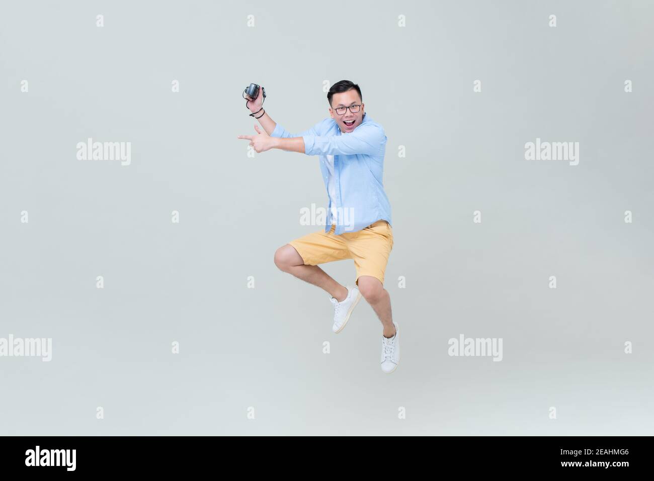 Young Asian man with camera jumping and pointing hand to empty space on light gray backgground Stock Photo