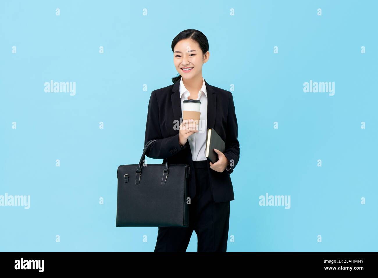 Professional Asian businesswoman in formal suit holding briefcase coffee cup and book isolated on light blue background Stock Photo