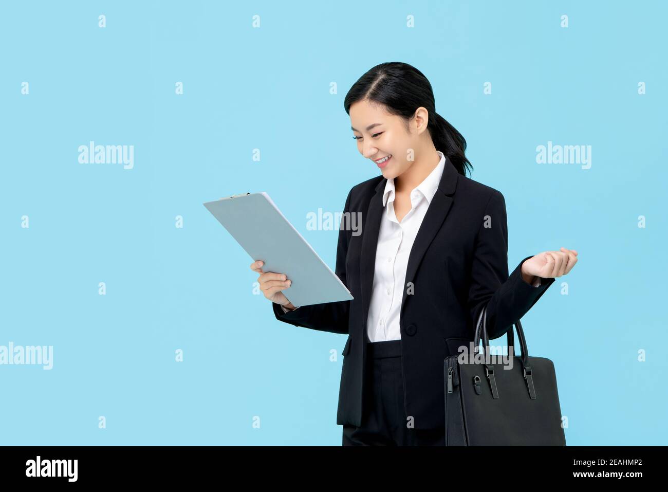 Professional Asian businesswoman in formal suit holding briefcase looking at clipboard isolated on light blue background Stock Photo