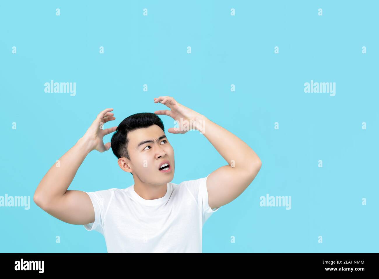 Upset angry Asian man with hands over head isolated on light blue background Stock Photo
