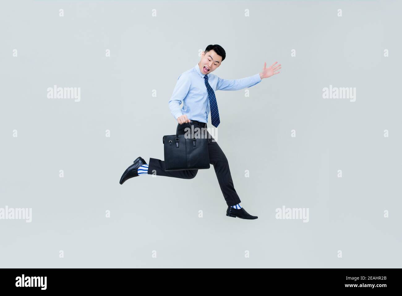 Full body of young happy smiling Asian professional man wearing business clothes and carrying briefcase jumping in midair isolated on light gray backg Stock Photo