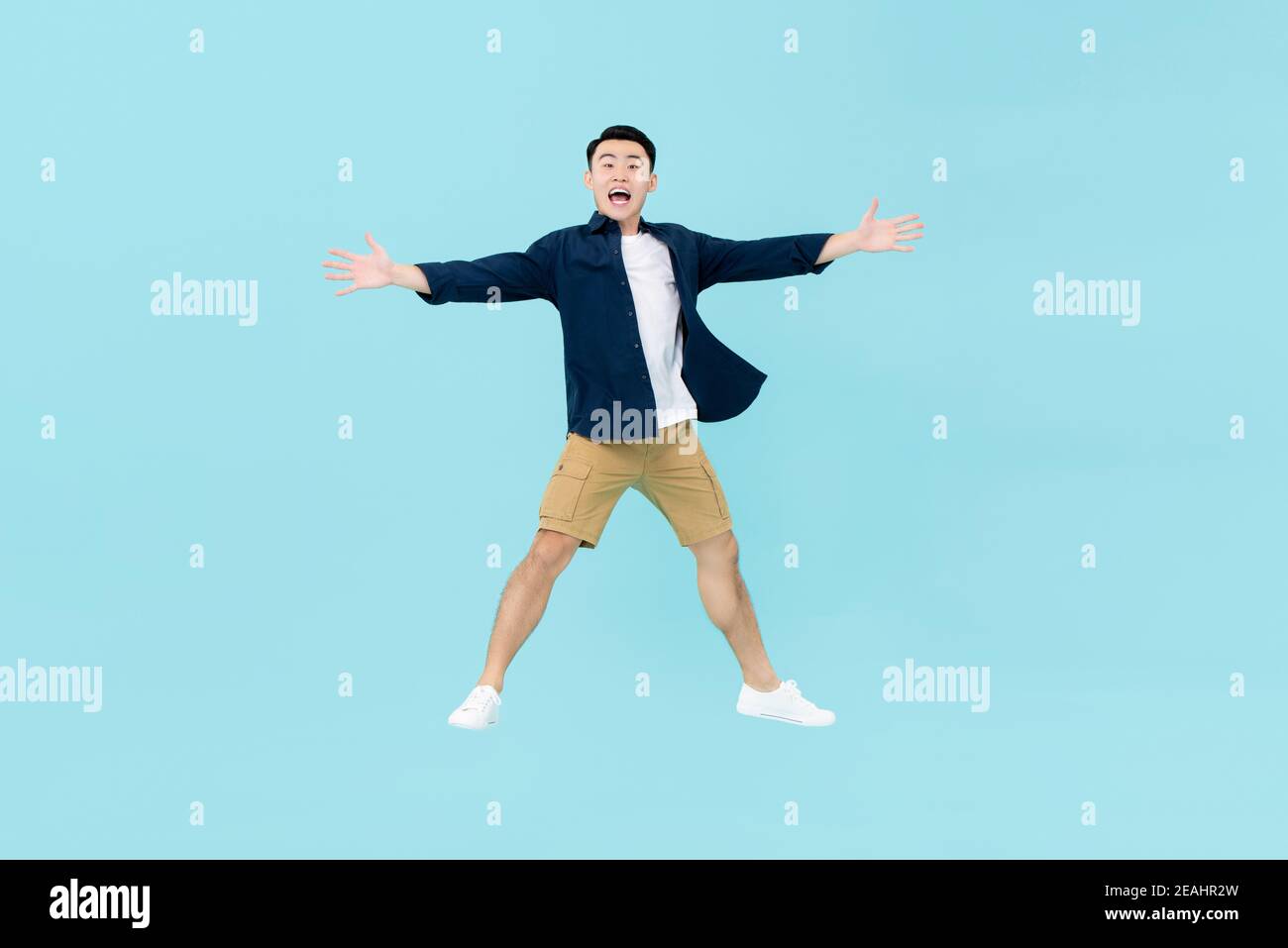 Full body of young dynamic handsome Asian man in casual clothes looking amazed and happy jumping in midair with arms and legs open isolated on light b Stock Photo