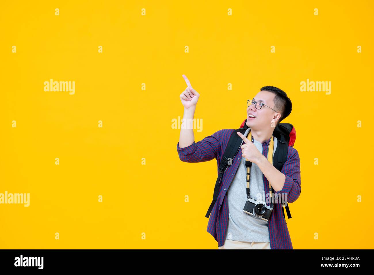 Young handsome happy Asian man in tourist clothes with backpack camera and glasses smiling and looking up while pointing with both hands beside copy s Stock Photo