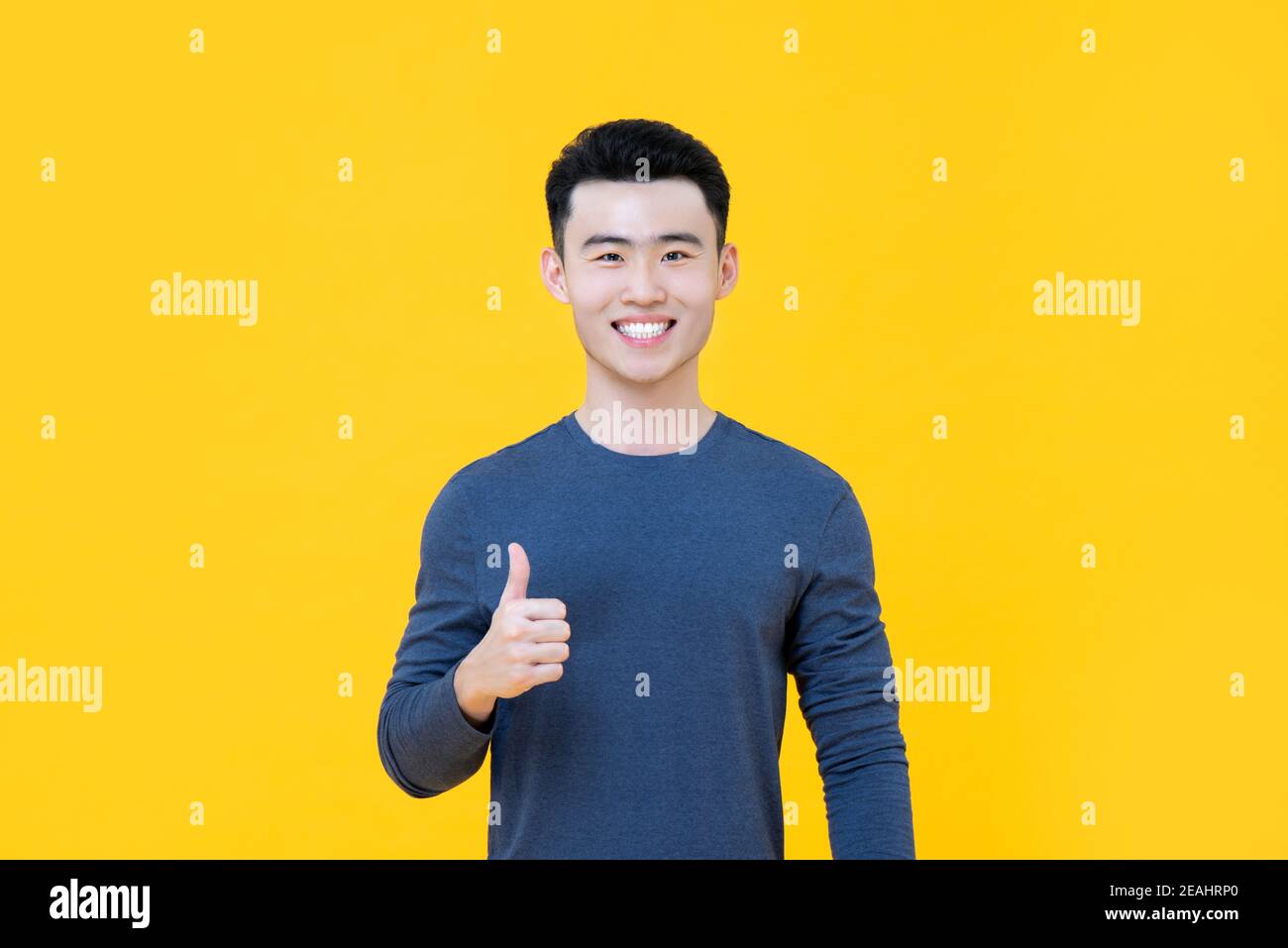 Young smiling Asian man giving thumbs up isolated on bright yellow studio background Stock Photo