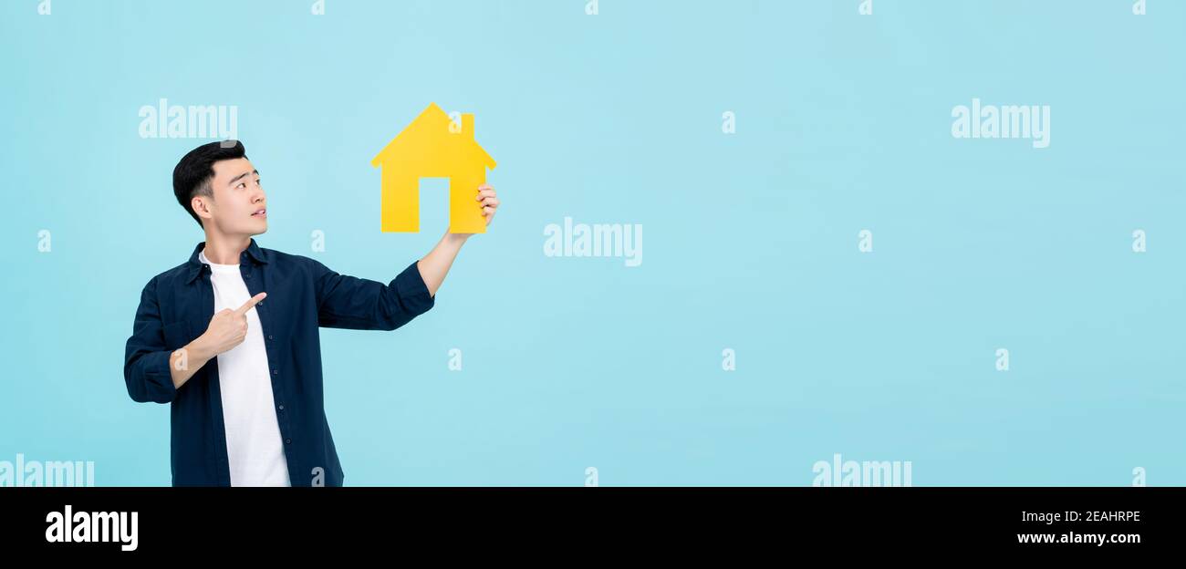 Young Asian man in casual clothes holding and pointing to house cut out isolated on light blue banner background for property concepts Stock Photo