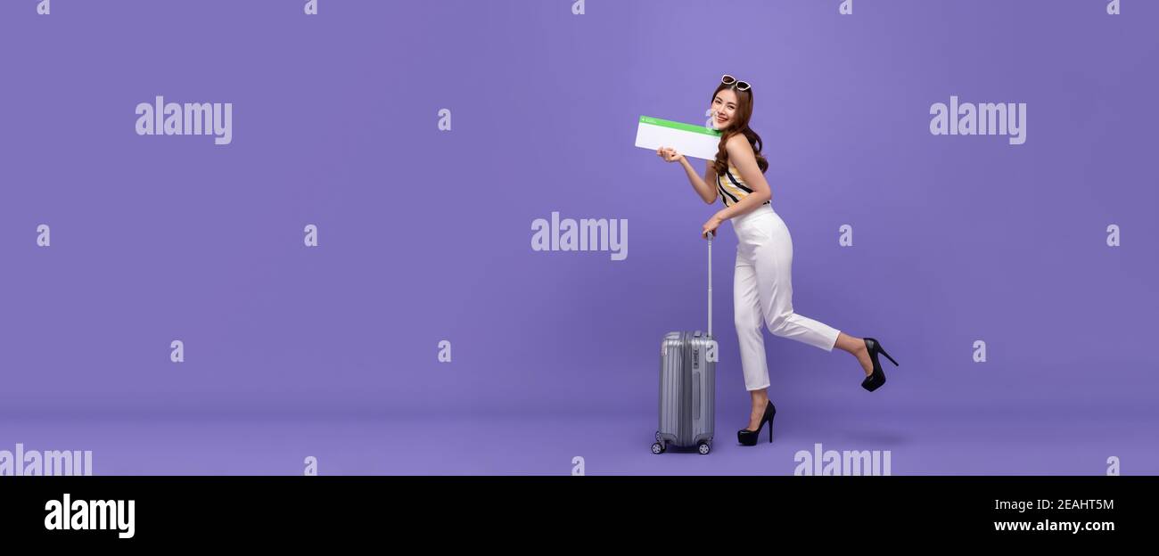 Young smiling Asian tourist girl with baggage showing airline boarding pass in colorful purple banner background with copy space Stock Photo