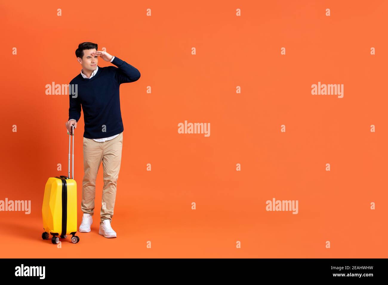 Full length of handsome young American tourist man holding baggage ready for travel looking away with hand on forehead isolated on orange background w Stock Photo