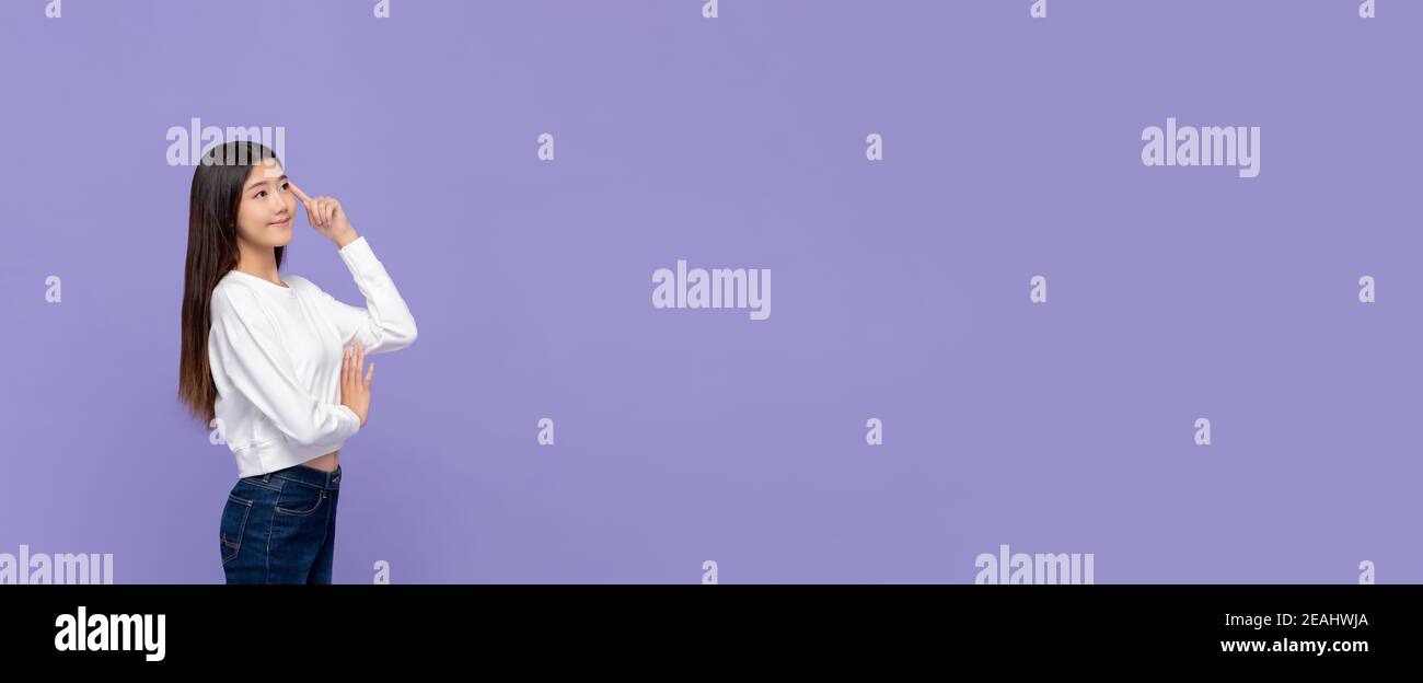 Attractive young smiling Asian woman thinking with hand touching head and looking away at empty space aside on purple banner  background Stock Photo