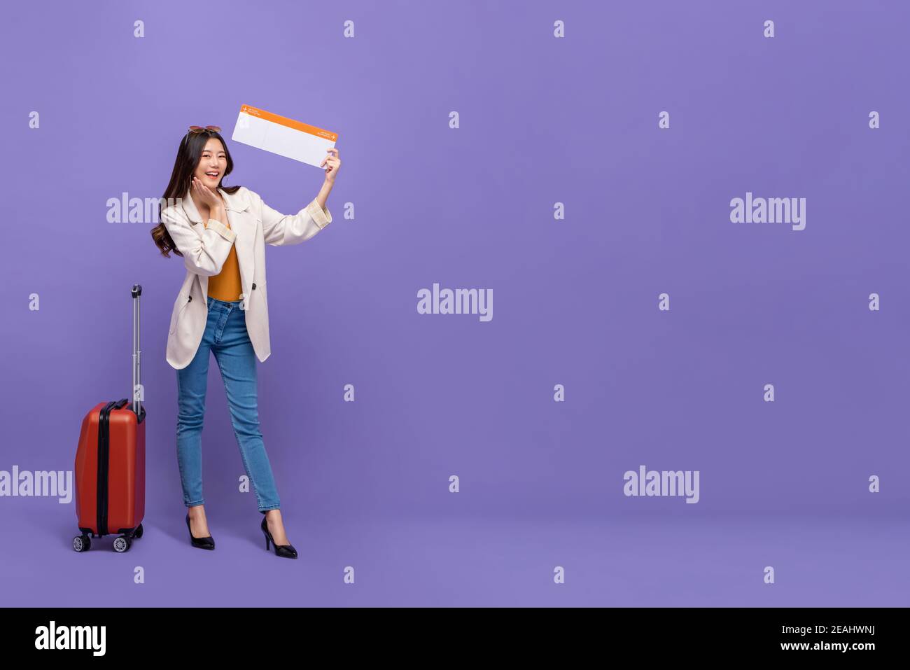 Full body of attractive young Asian woman tourist with luggage showing confirmed boarding pass isolated on purple background with copy space Stock Photo