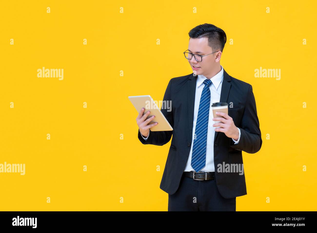 Focused Asian businessman looking at tablet computer while drinking coffee isolated on yellow background Stock Photo