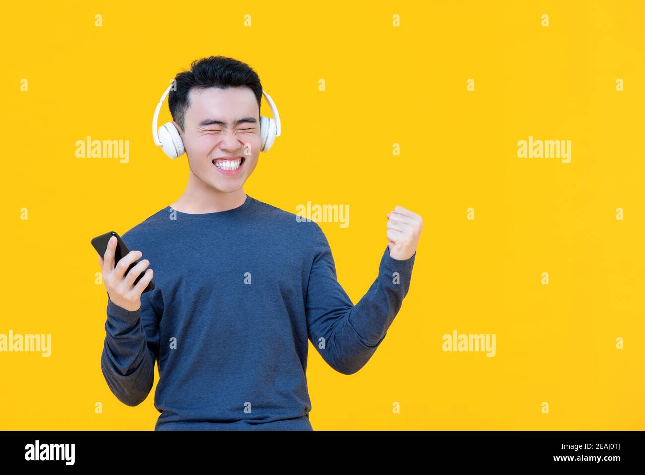 Amazed Asian man wearing headphones listening to his favourite songs smiling and clenching his fist isolated on yellow studio background Stock Photo