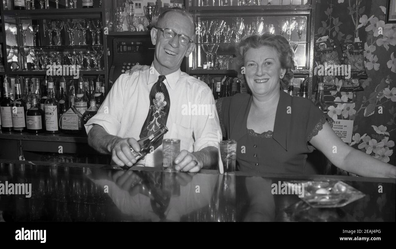 1940s, historical, bar or saloon owner with his wife at his bar pouring a drink, Oregon, USA. Behind them bottles of the leading spirit brands and glasses. Stock Photo