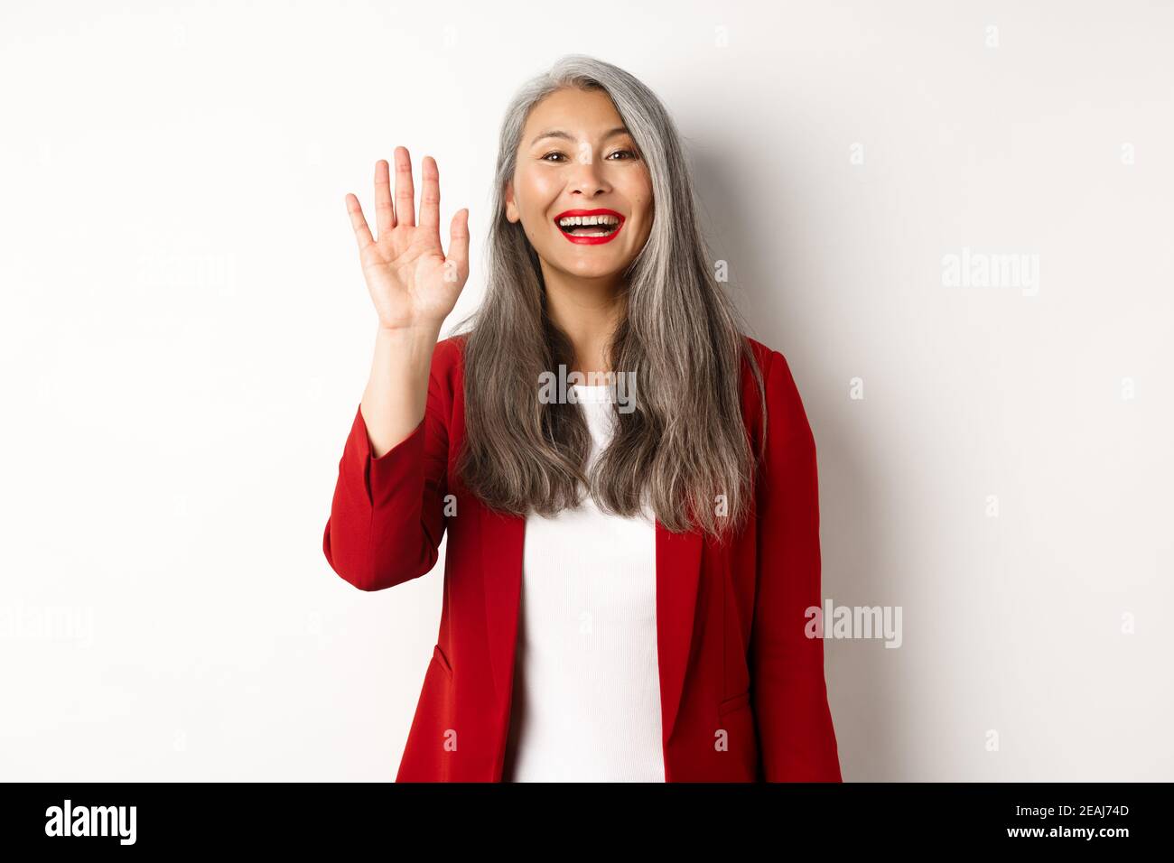 Friendly asian senior lady in elegant blazer saying hi, waving hand and greeting you with happy smile, standing over white background Stock Photo