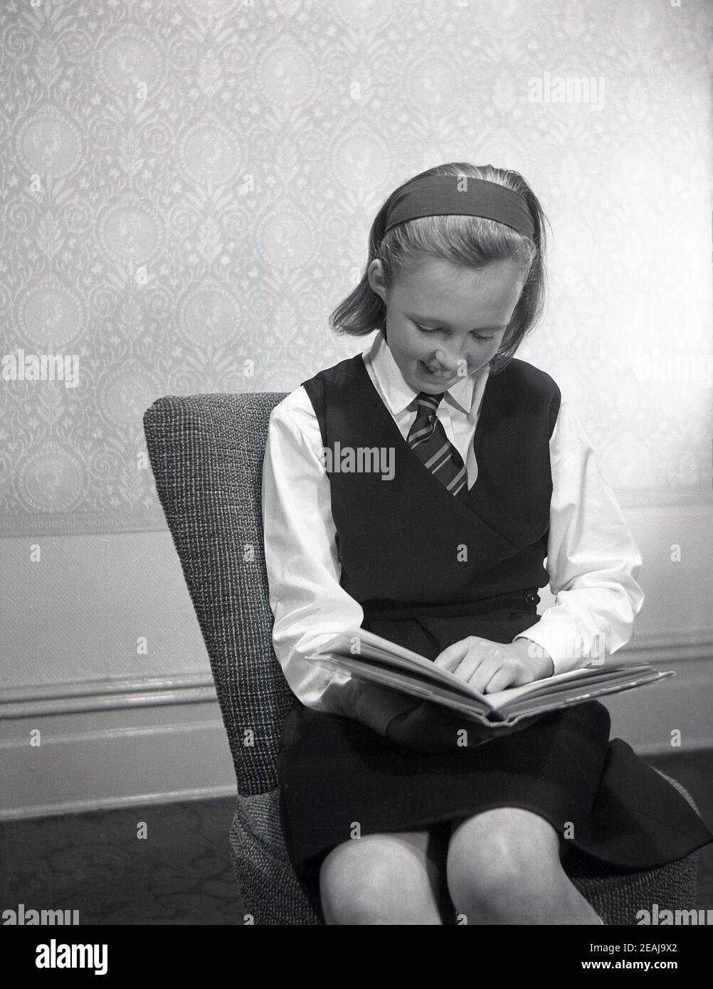 1950s, historical, a teenage schoolgirl in her uniform sitting in a room on a small chair reading or studying a book, England, UK. Stock Photo