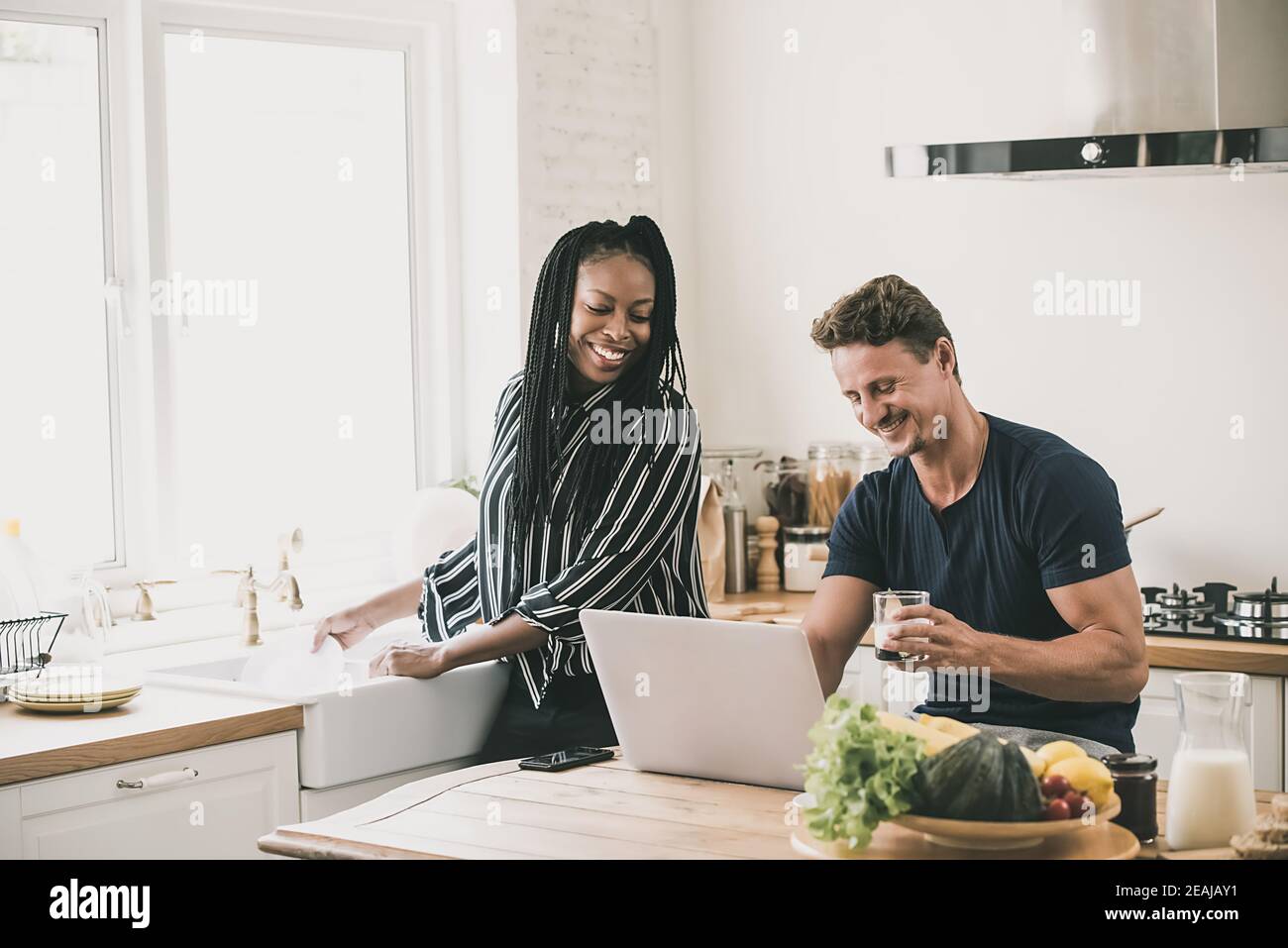 Happy married interracial couple looking at laptop computer together while doing daily moring routines in the kitchen at home Stock Photo