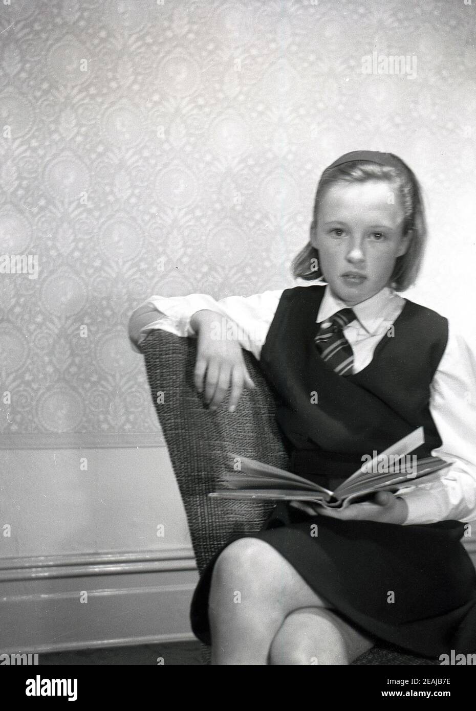 1950s, historical, a teenage schoolgirl in her uniform sitting in a room on a small chair with a school book, England, UK. Stock Photo
