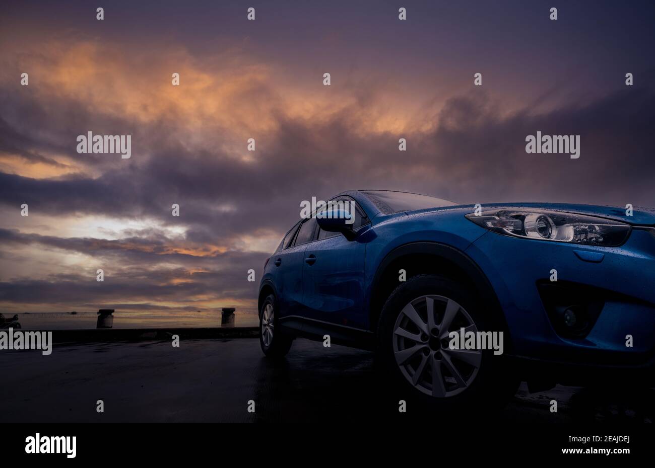 Luxury SUV car with raindrops. Front view new blue SUV car parked on concrete parking lot near sea beach with sunset sky. Electric vehicle concept. Road trip travel. Car covered with water drops. Stock Photo
