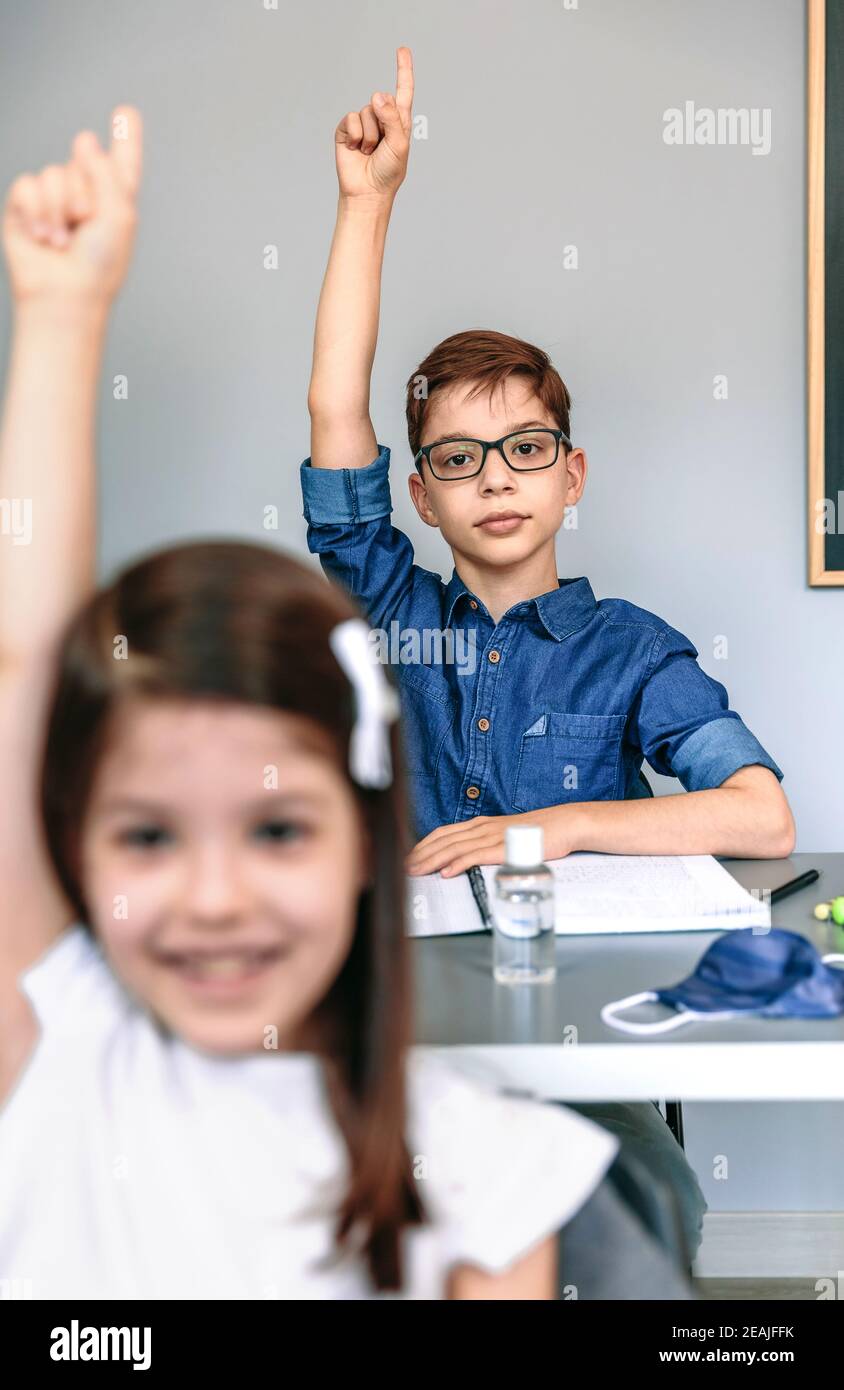 Students with mask on table raising hands at school Stock Photo