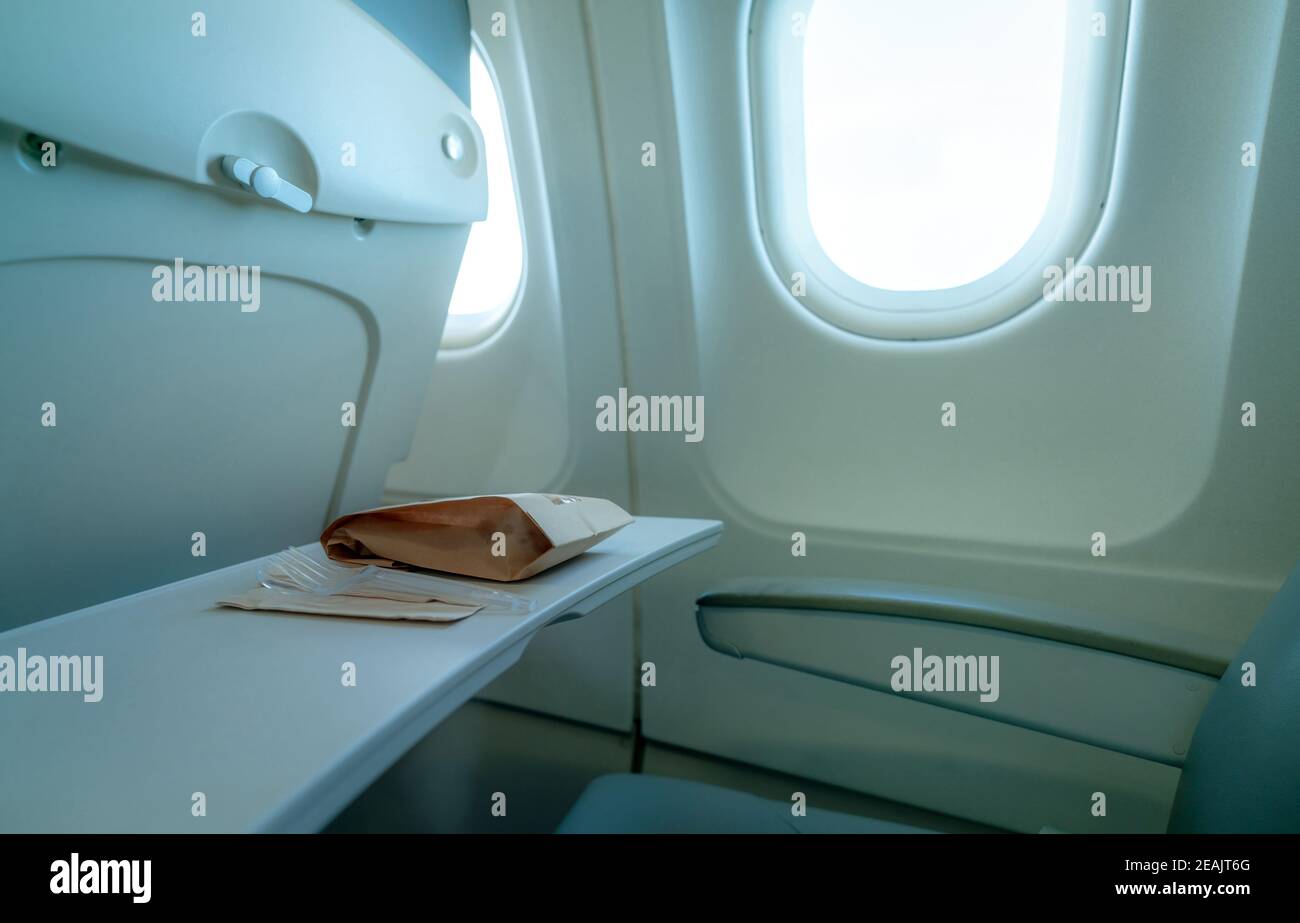 Plane window. Food in brown paper bag and plastic fork and spoon on plastic airplane tray table at seat back. Economy class airplane window. Inside of commercial airline. Seat with armchair. Stock Photo