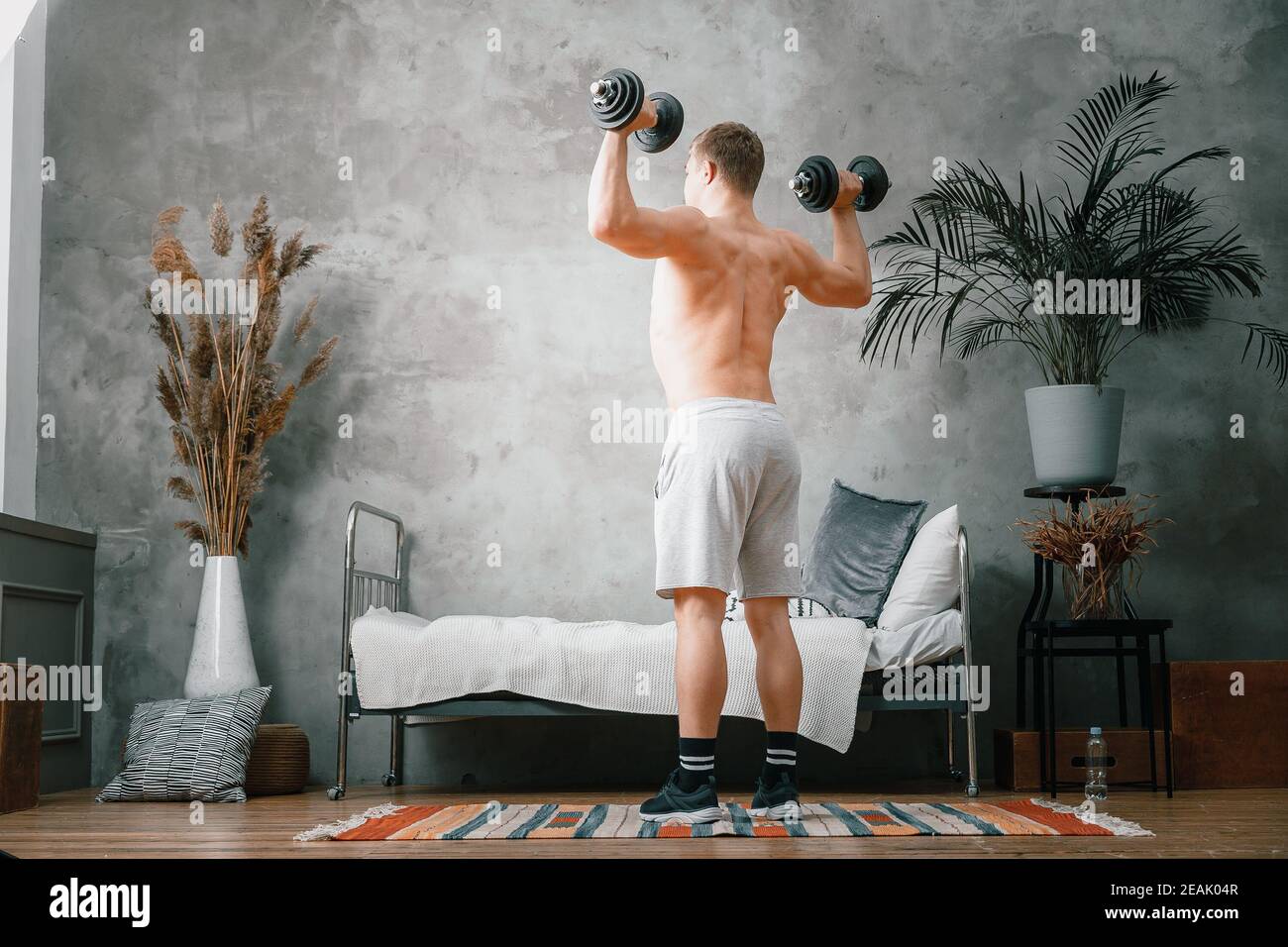 A athlete with blond hair shakes biceps in dumbbells in the bedroom, online training. The young