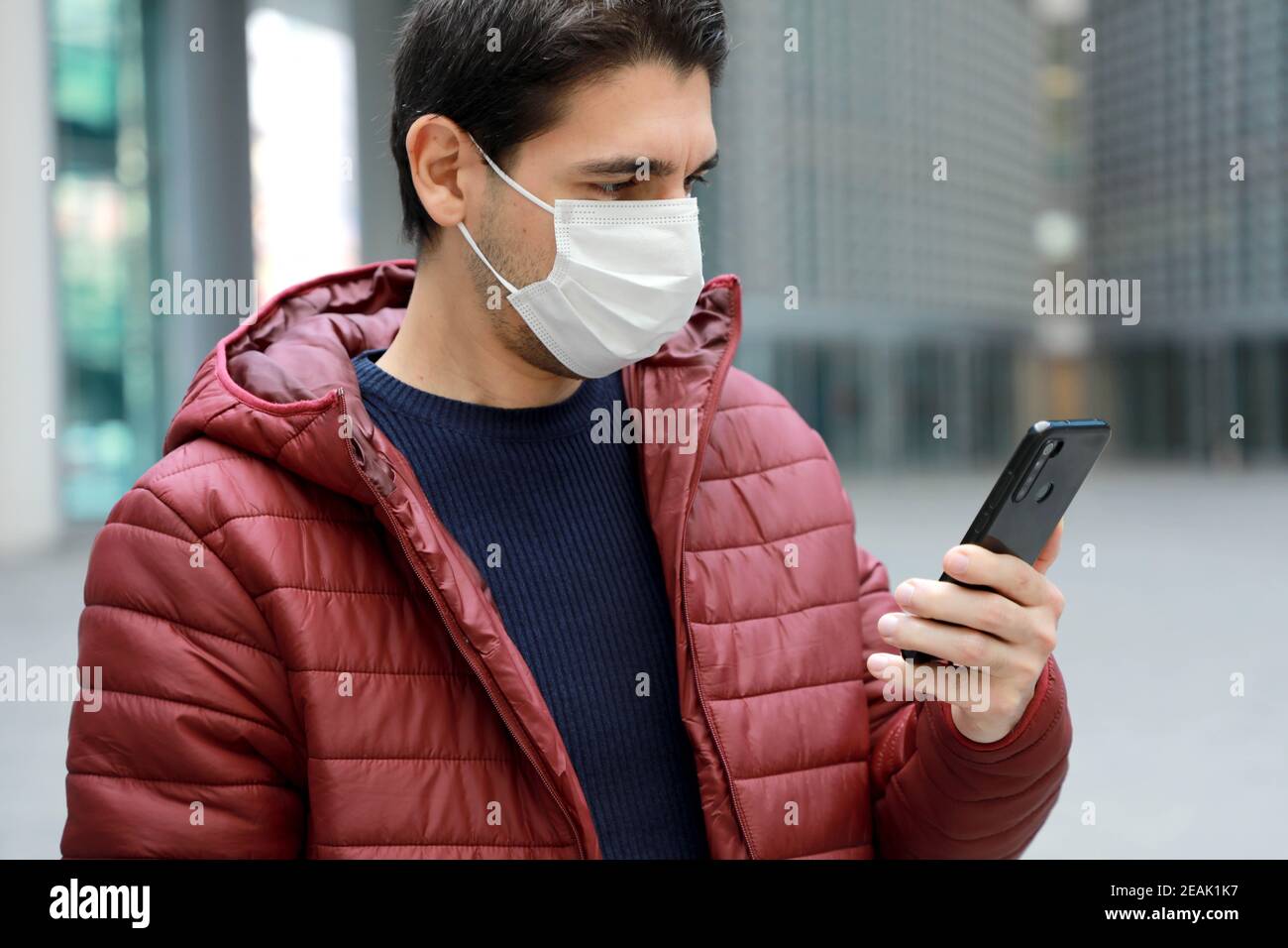 COVID-19 Portrait of young man wearing protective mask reading news on smartphone app in modern city street Stock Photo