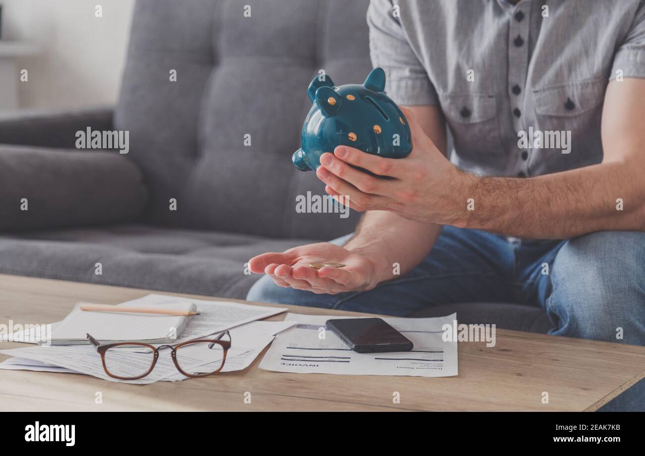 Man sitting at a table full of unpaid bills shakes out the last penny from the piggy bank. Spend your last savings. Unemployment, poverty, bankruptcy concept. Stock Photo
