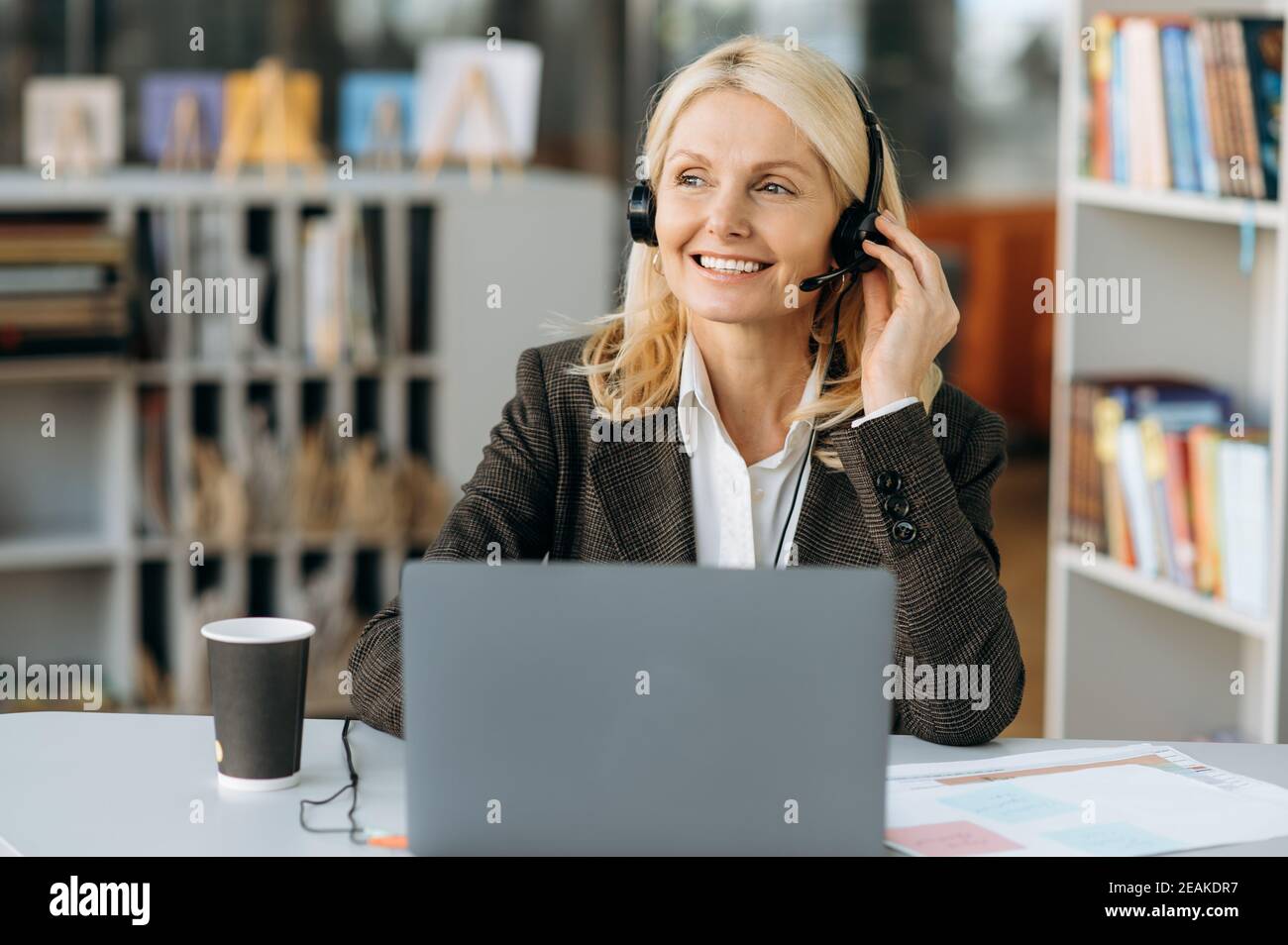 Satisfied female employee in headset and stylish formal suit sits at the desk and friendly smiling. Elegant senior blonde business woman works in support service, consulting clients, communication Stock Photo