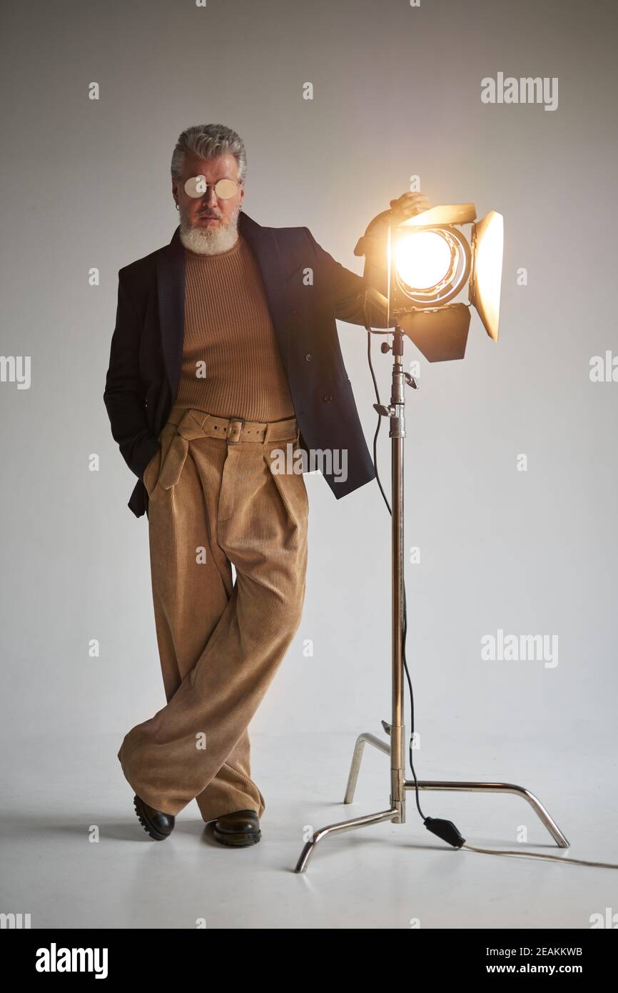 Full length shot of fashionable grey haired middle aged man in glasses looking at camera, standing next to studio spotlight while posing over white background. Fashion photoshoot, style concept Stock Photo