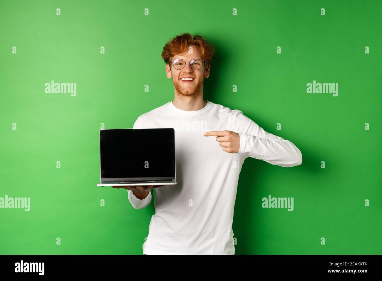 Happy redhead guy in glasses and white long-sleeve t-shirt, pointing finger at blank laptop screen and smiling, standing over green background Stock Photo
