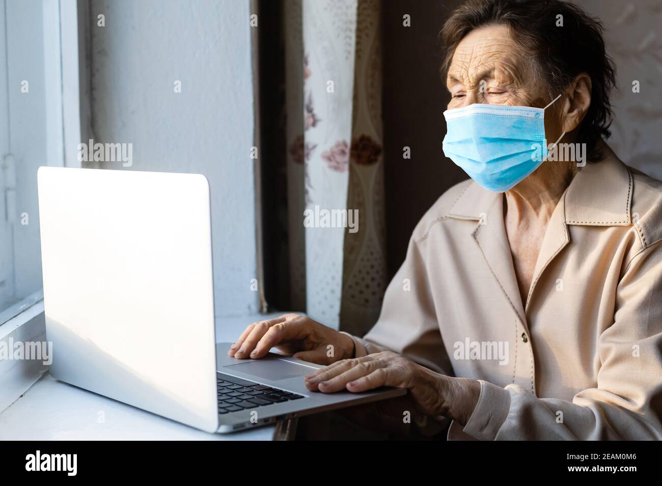 an elderly woman in a mask near the window with a laptop Stock Photo