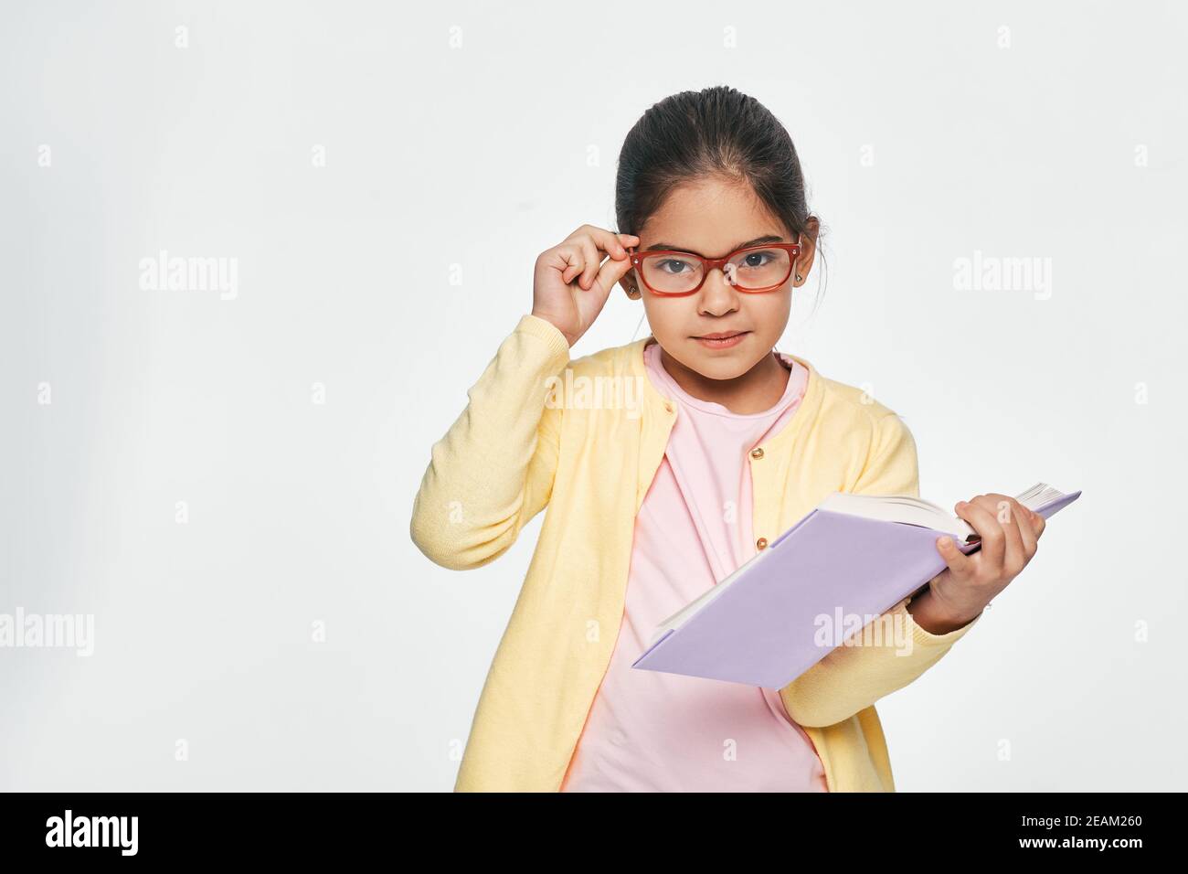 Mixed race little girl wearing eyeglasses holds book looking pensive at camera, on light grey background Stock Photo