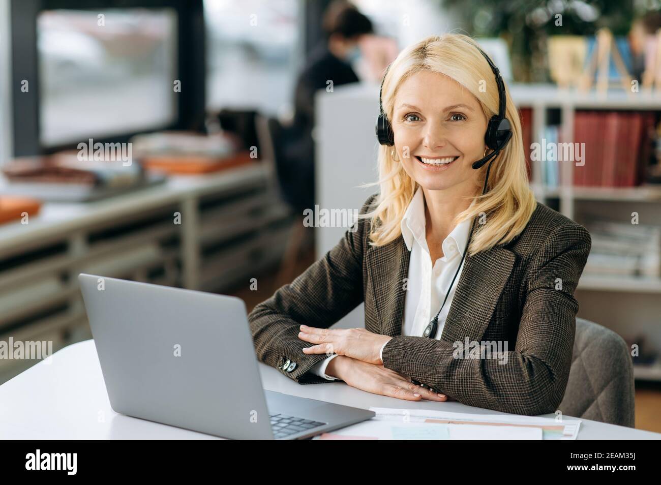 Joyful elegant female operator of call center in headset is looking at the camera, smiling. Intelligent business woman is sitting at the table in office, working in support service, consulting people Stock Photo