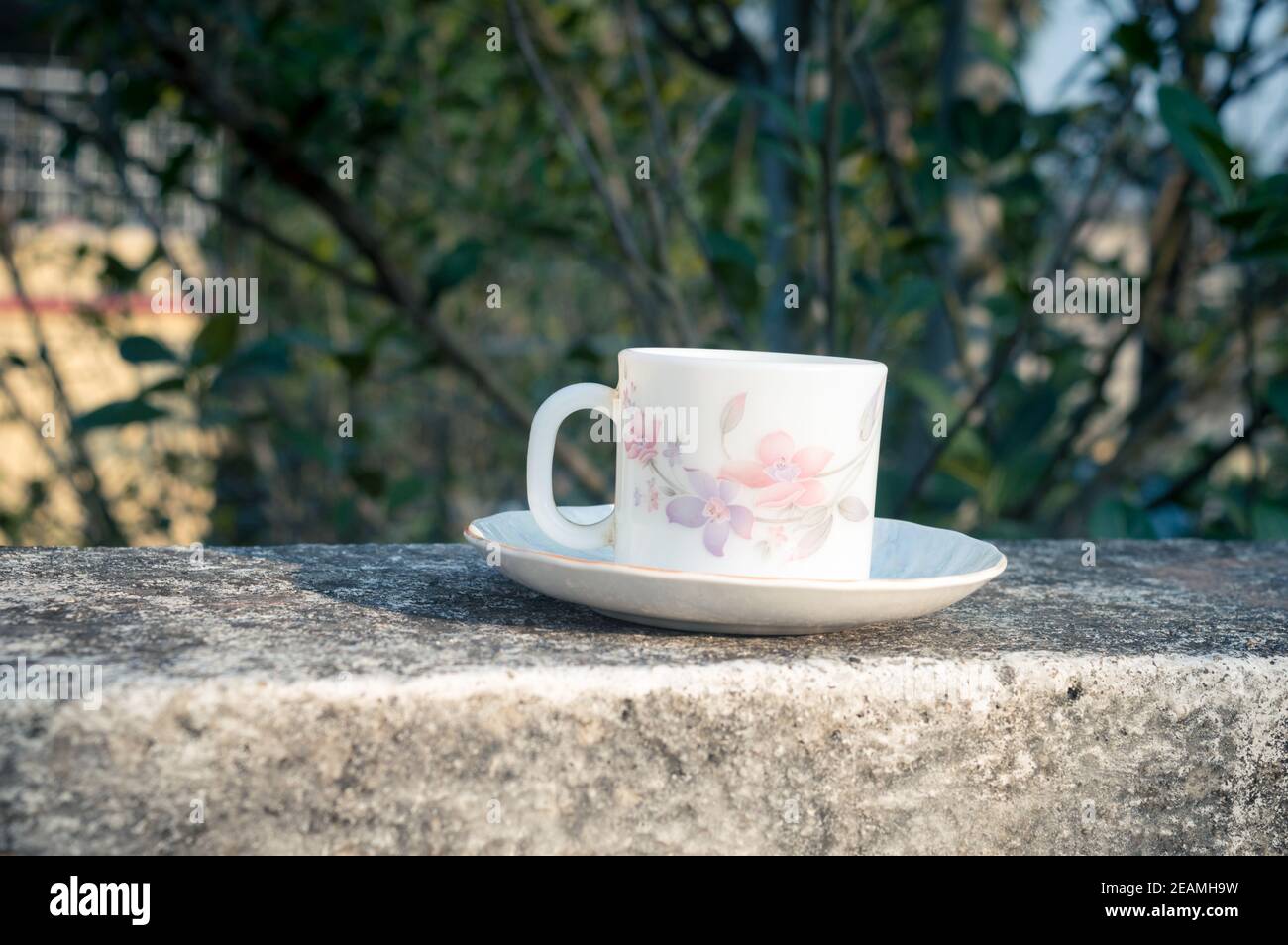 Coffee cup in morning sunlight. Summer fresh cool look. White coffee cup on saucer for hot drink on roof beam of a residential building with bokeh background. Stock Photo
