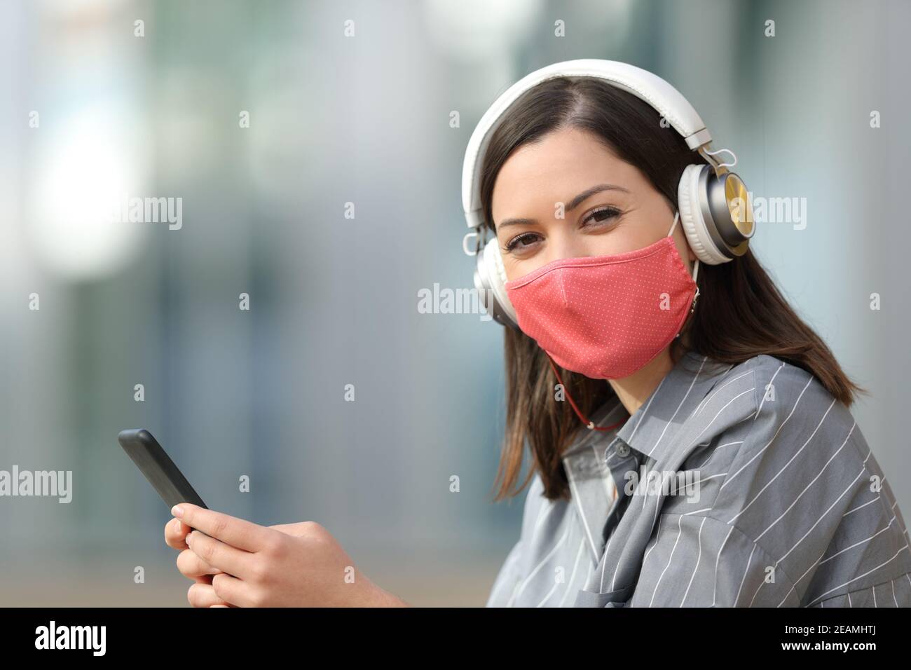 Happy woman with mask and headphones holding phone Stock Photo