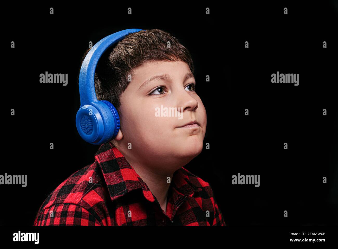 Caucasian white boy teenager in a stylish plaid shirt in red listening to music in big blue wireless over-ear headphones on isolated black background Stock Photo
