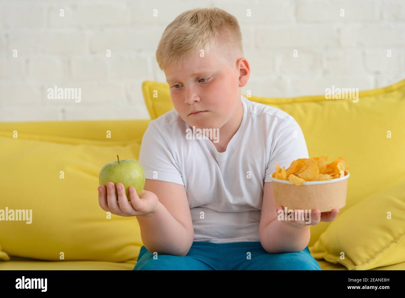 Sad fat boy looking at an apple and holding a cup of chips in another hand Stock Photo