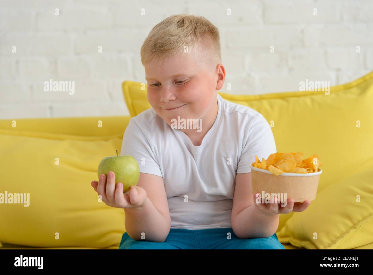 Happy fat boy looking at an apple and holding a cup of chips Stock Photo