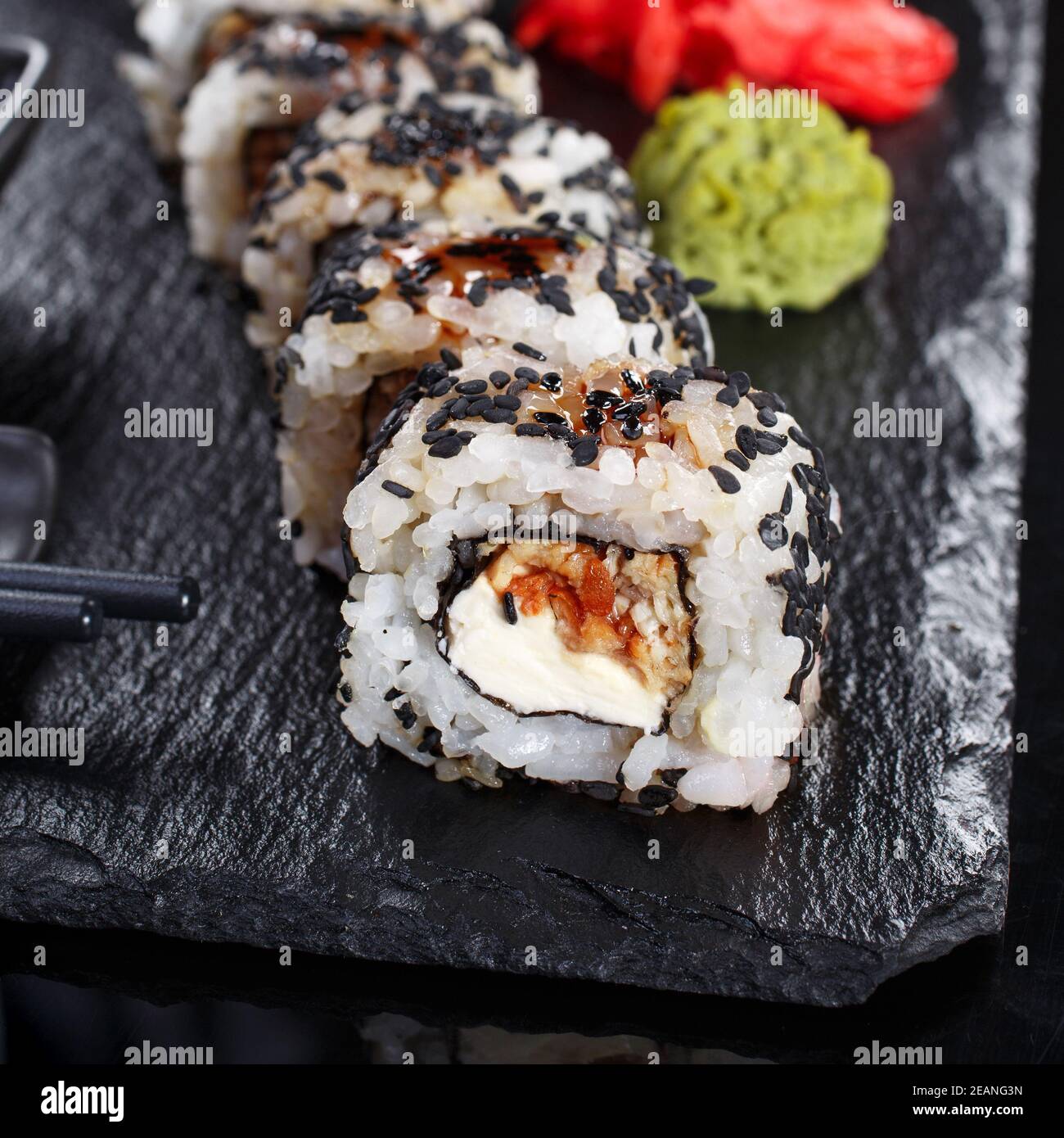 Japanese Traditional Cuisine. Sushi roll with sesame seeds is served with chopsticks and soy sauce on black slate, decorated with wasabi and ginger. Stock Photo