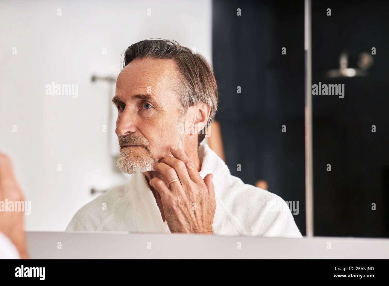 Senior man touching her face in bathroom in from of a mirror. Male in bathrobe looking at his reflection  Stock Photo